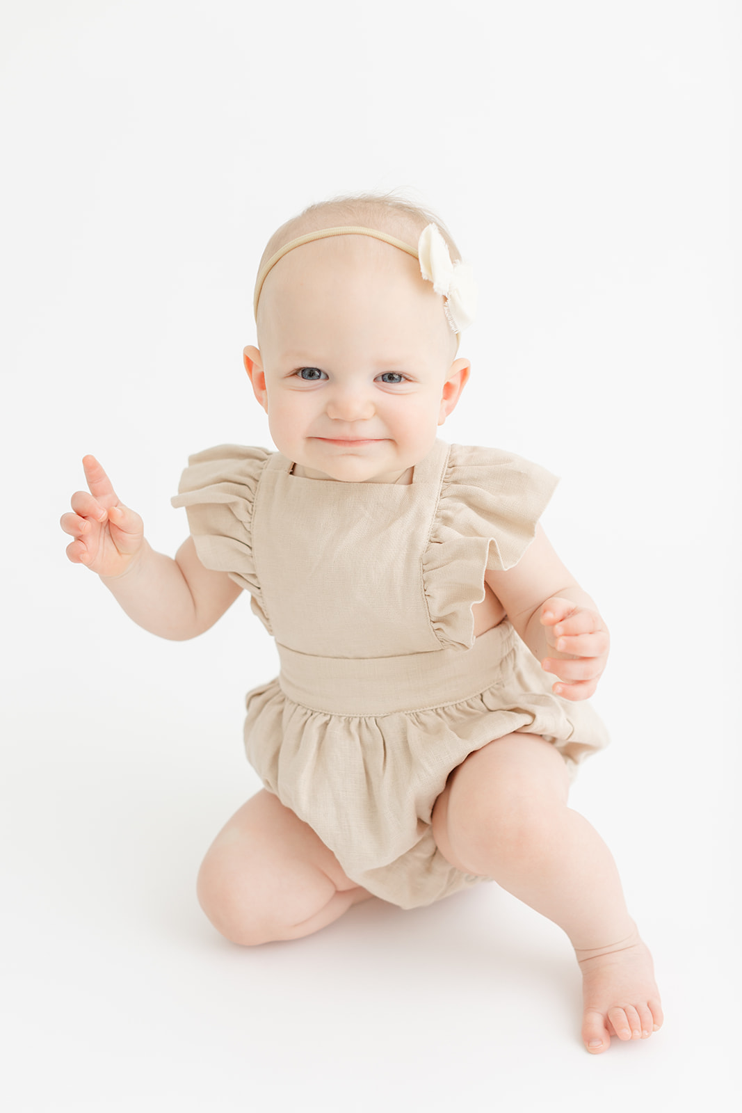 A young baby smiles while playing in a studio in a cream dress and bow