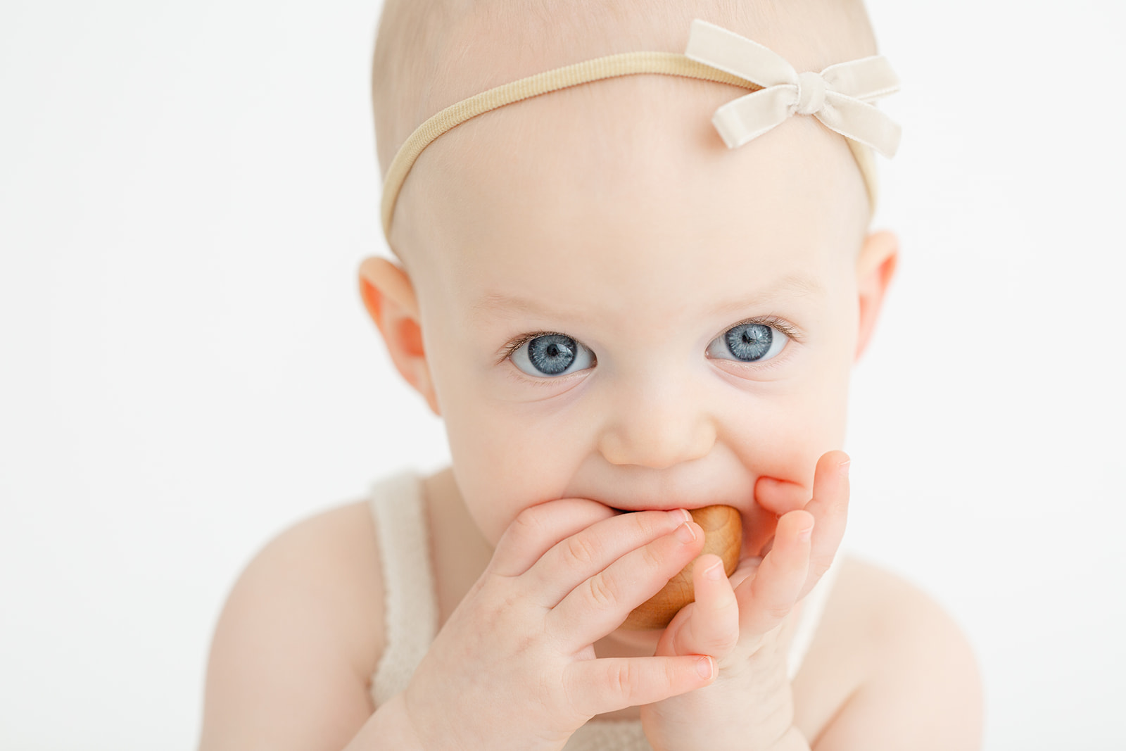 A young baby with blue eyes chews on a wooden toy while wearing a headband bow after using Unfurling Birth