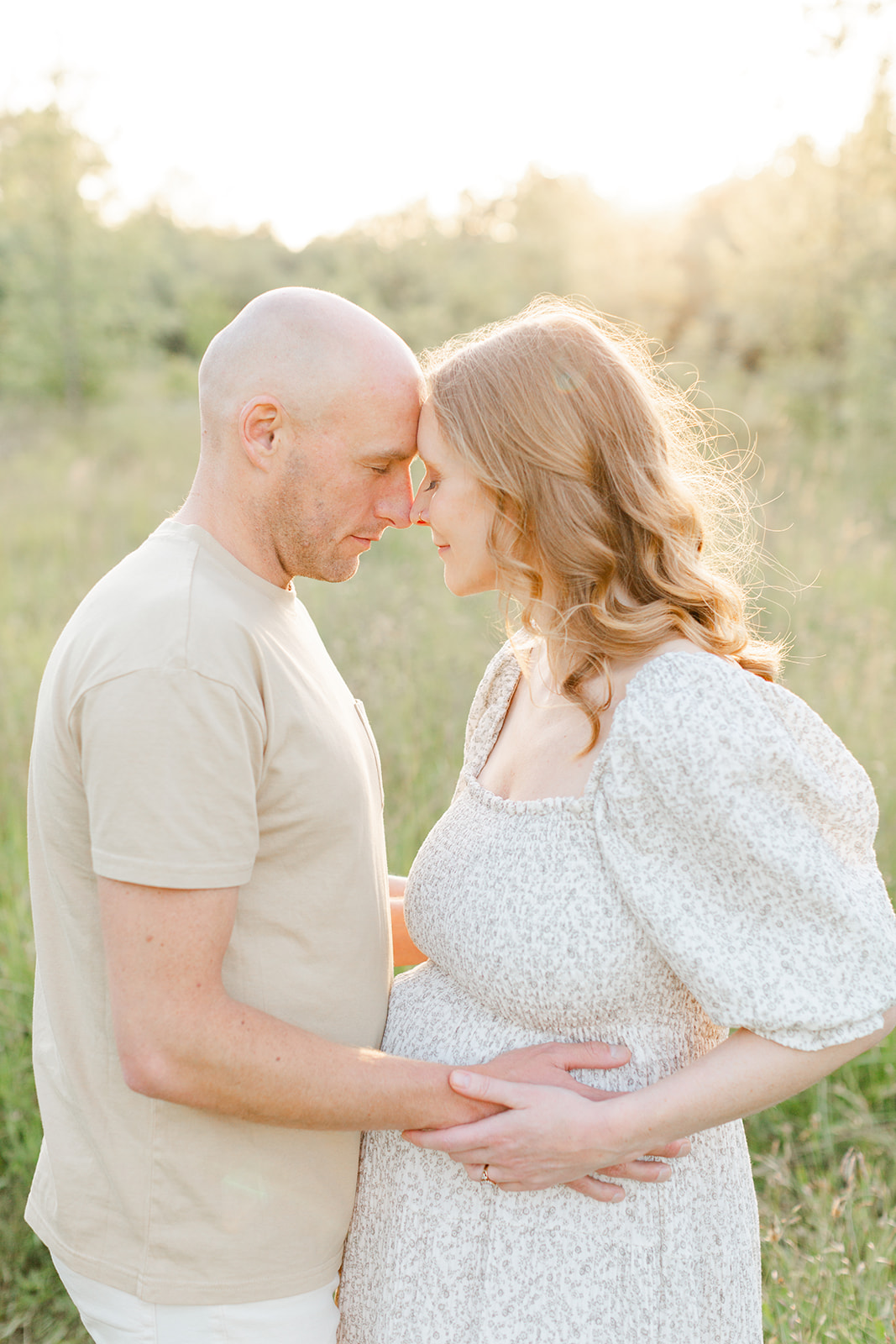 Expecting parents hold the bump and touch foreheads while standing in a field of tall grass at sunset after visiting Doula Love Portland