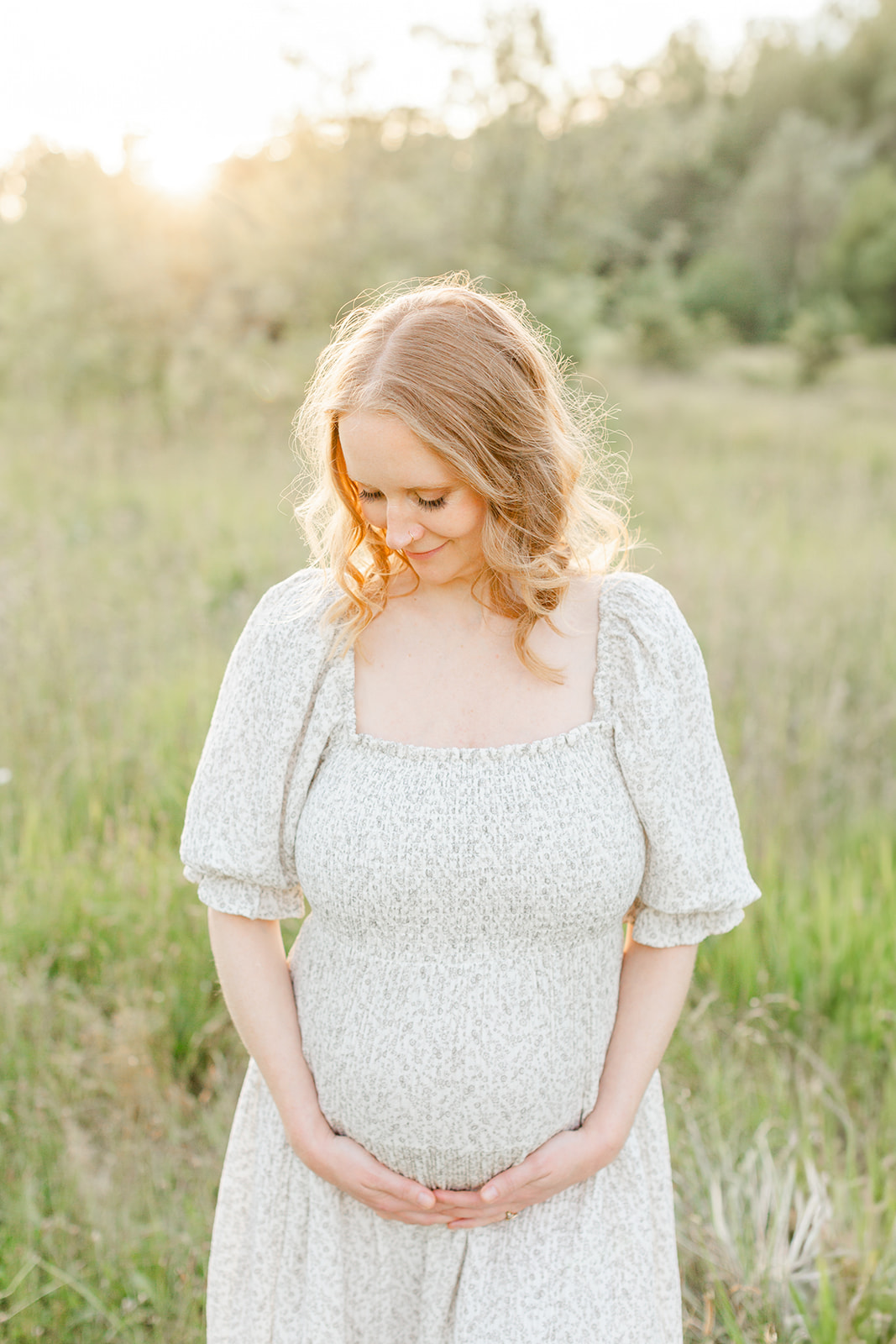 A mother to be in a floral print dress smiles down to her bump in a field of tall grass at sunset after meeting Doula Love Portland