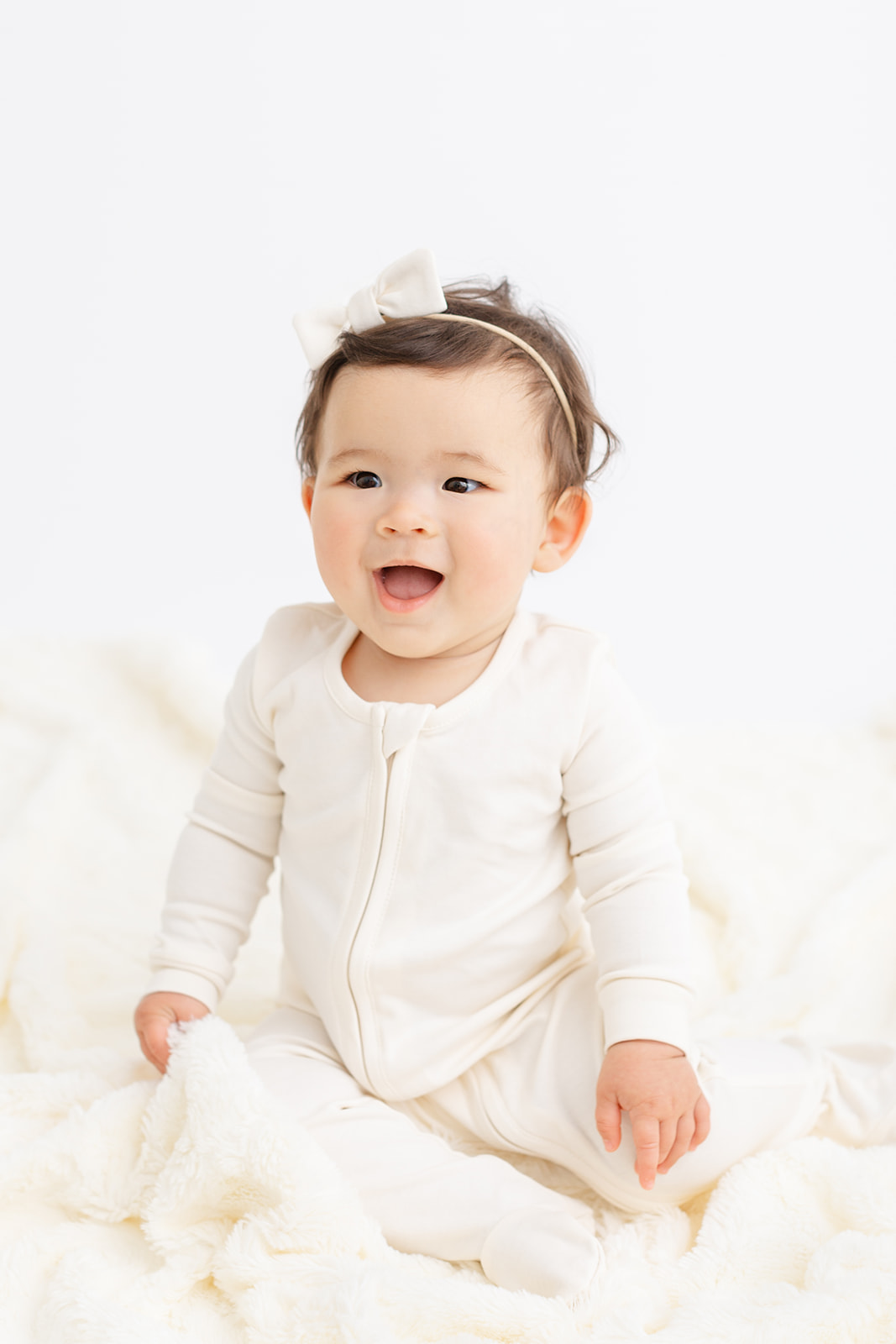 A young toddler in a white onesie smiles big while sitting on a white blanket in a studio