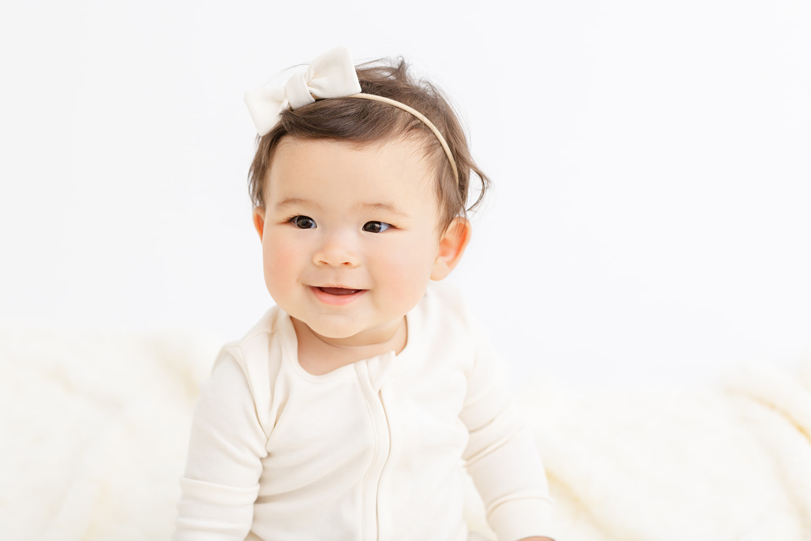 A smiling toddler girl sits in a studio with a white bow and matching pajamas