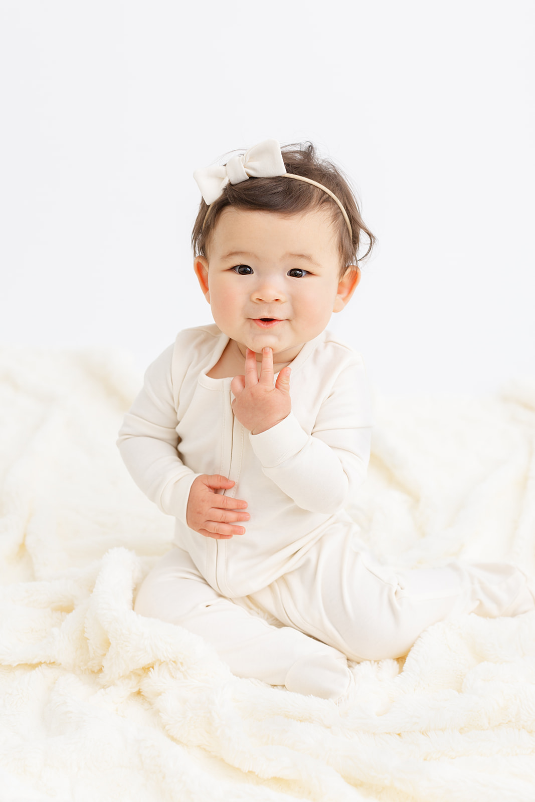 A smiling toddler girl in white pajamas sits on a white blanket in a studio with fingers on her chin before visiting Ready Set Grow