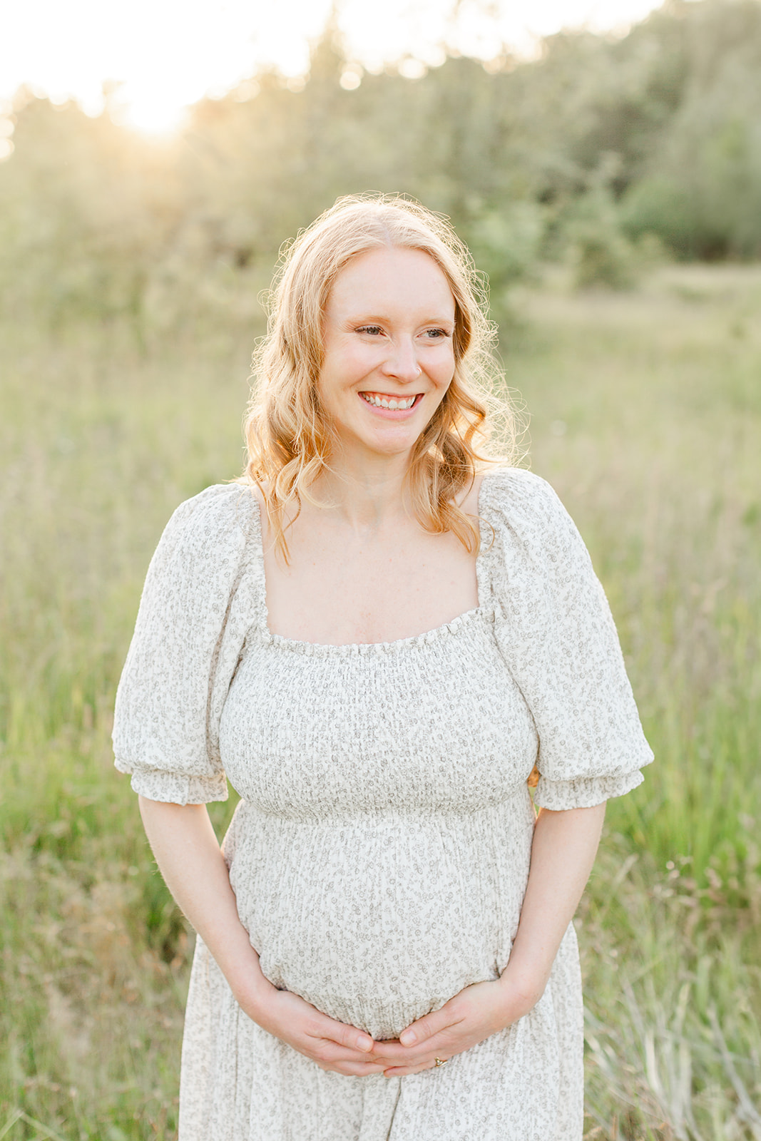 A smiling pregnant woman holds her bump while walking through tall grass at sunset