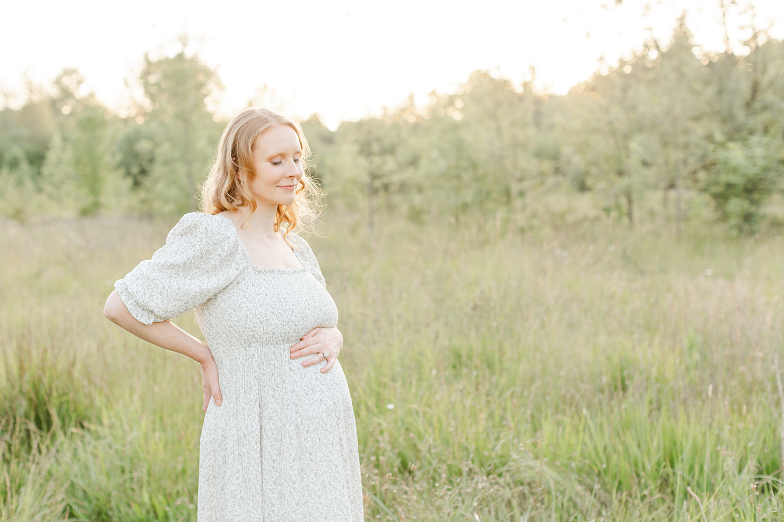 A happy mother to be stands in a pattern maternity dress in a field of tall grass at sunset after meeting Bliss Lactation