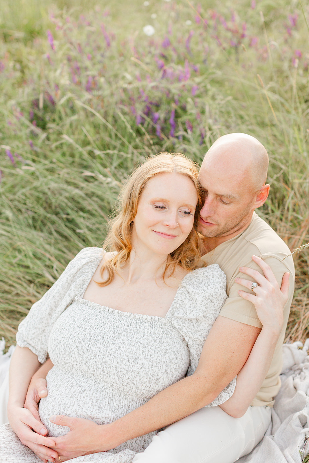 A mom to be leans into the lap of her husband while sitting on a picnic blanket in tall grass after meeting Bliss Lactation