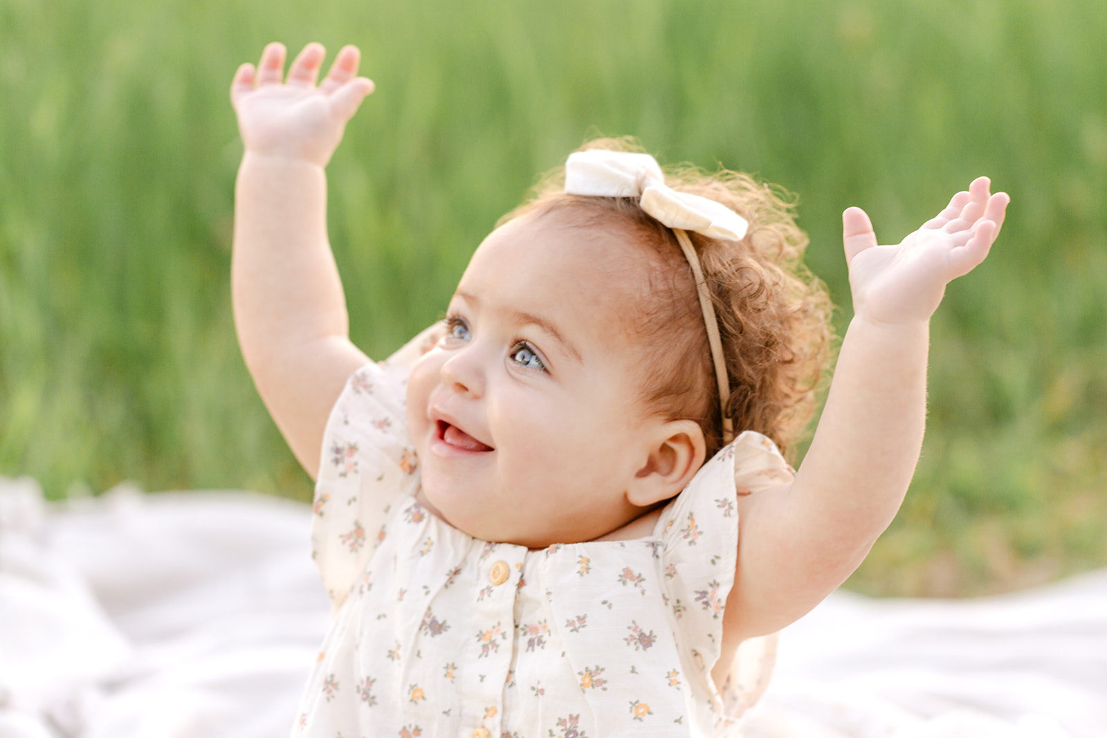 A young toddler girl smiles and raises her hands while playing on a picnic blanket in a park after meeting Luna Lactation