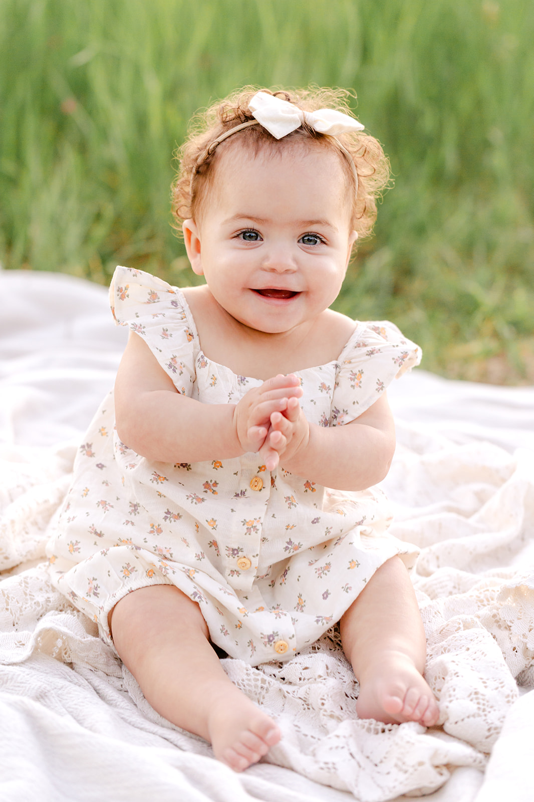 An infant girl in a dress sits on a blanket in a park lawn clapping with a smile after meeting Luna Lactation