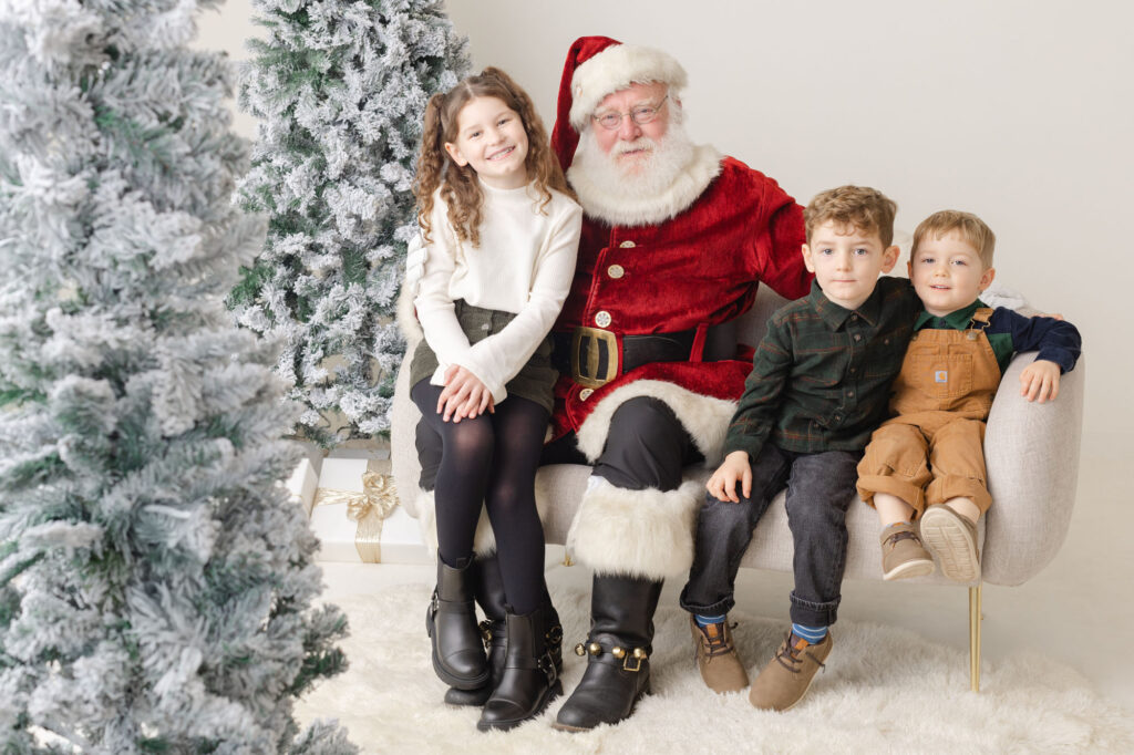 Three kids sitting on a couch with Santa looking and smiling at the camera. They attending Ashlie Behm Photography's Santa Studio sessions event.