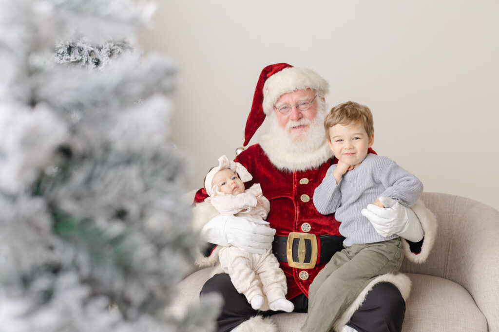 Santa holding a baby and a little boy on his lap and they are smiling and looking at the camera. 