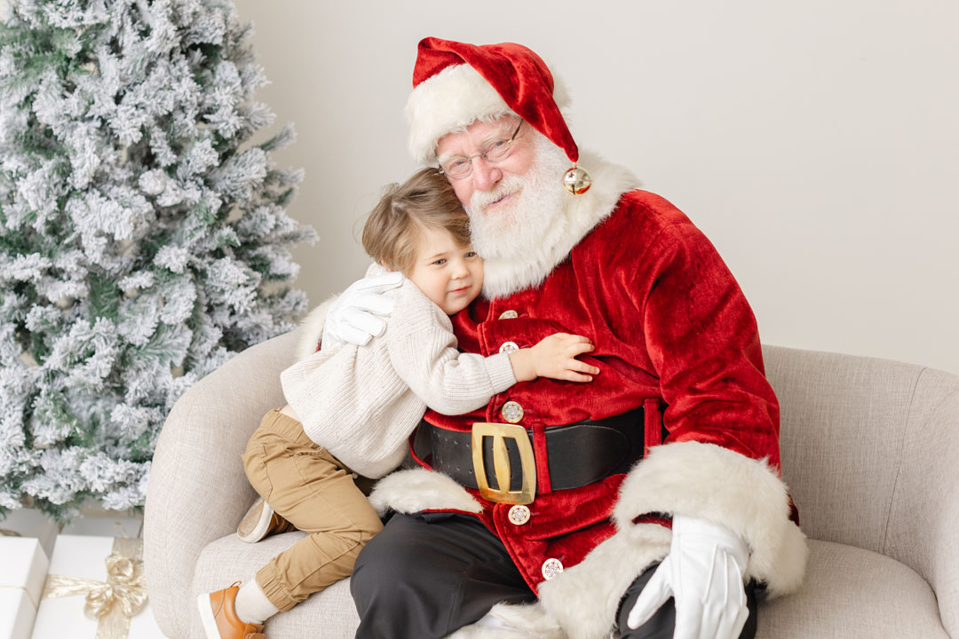 Little boy in neutral clothes is hugging Santa with a christmas tree behind them.