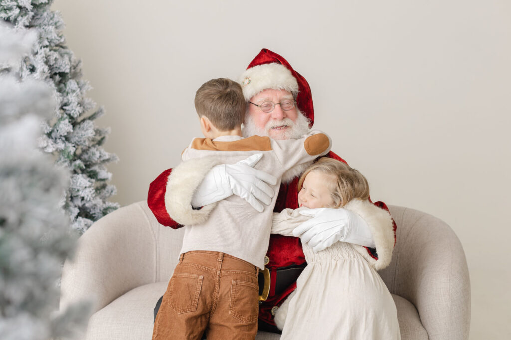 Two little kids are hugging Santa at their Santa Studio Session with Ashlie Behm Photography
