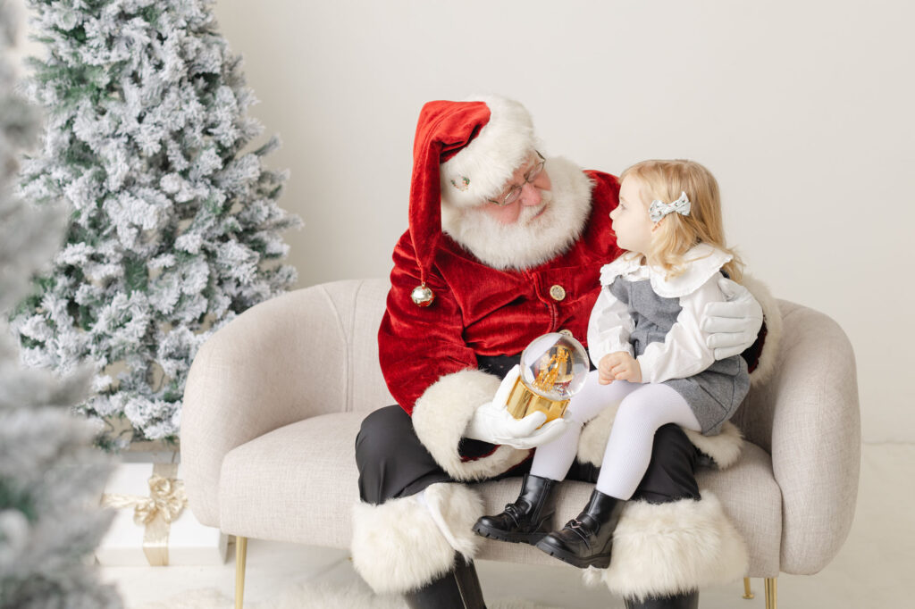 Portland Santa photos with Ashlie Behm Photography. Little girl sitting on Santa's lap looking at him while he holds a snowglobe.