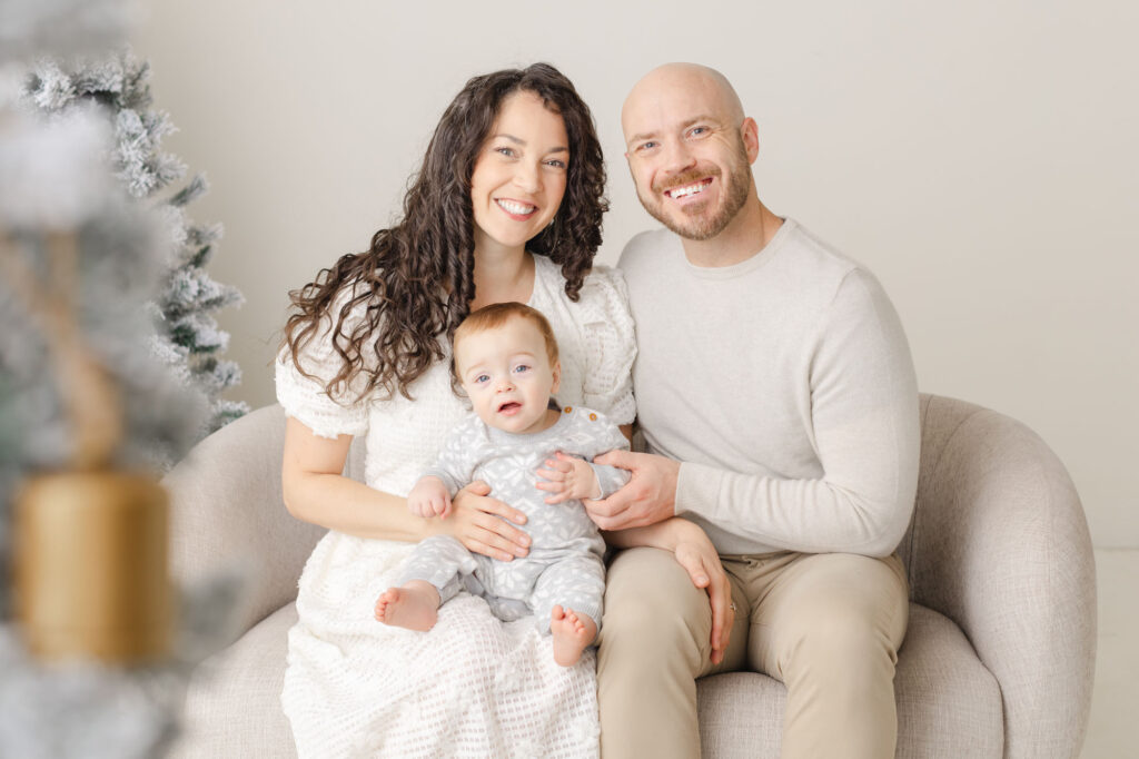 Mom, Dad and little baby sit on a couch near a christmas tree for their annual holiday family portrait.