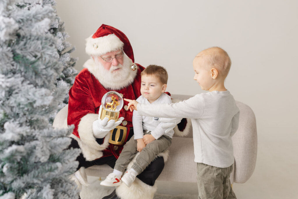 Santa studio sessions in Portland, Oregon Two boys with Santa one is on his lap while he holds a snowglobe and the other boy is standing nearby pointing at the snowglobe.