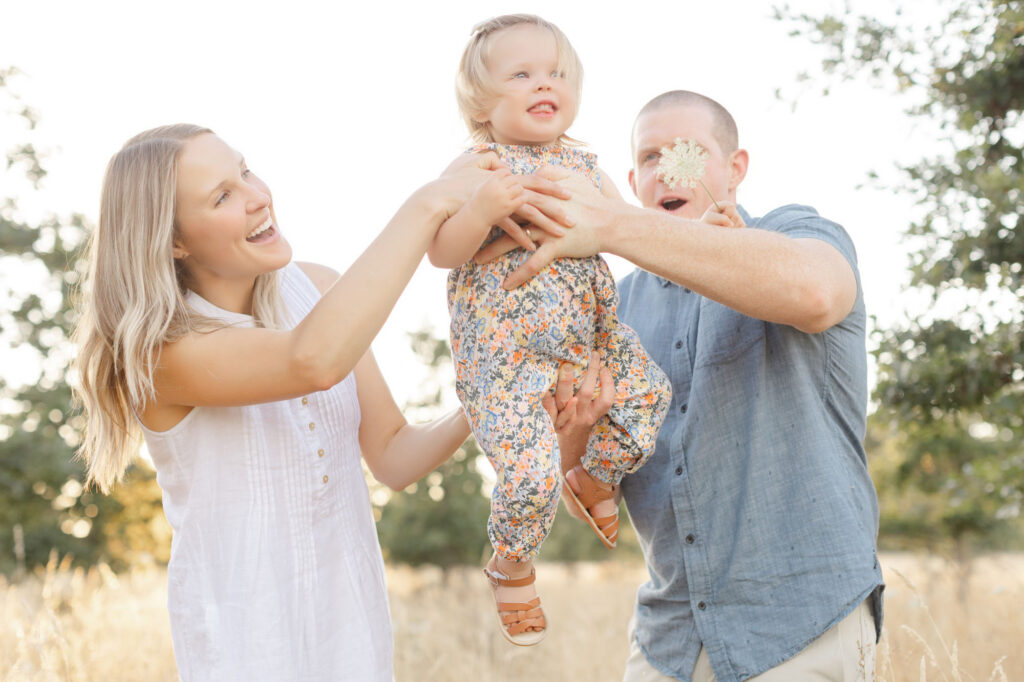 Family of three with parents and a toddler outdoor for summer family photos at Graham Oaks Nature Park with Ashlie Behm Photography. The family is wearing light colored outfits with the toddler in a cute floral jumpsuit. They are all smiling as the parents hold the toddler between them and lift her up in the air.