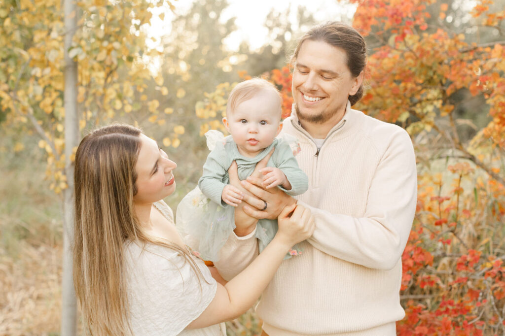 Family photo of mom, dad and baby are all dressed in creams and greens. Mom and Dad are holding baby between them and looking at her and smiling. Baby is looking at the camera. They have some beautiful fall foliage behind them.