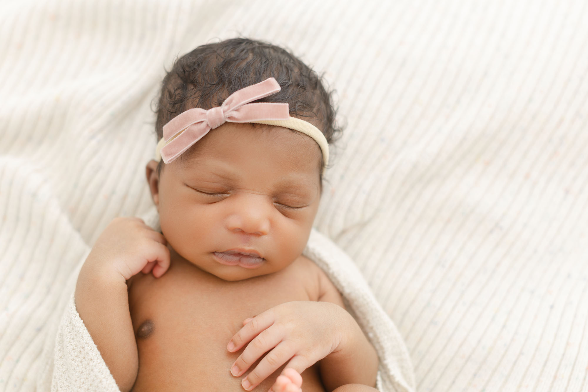 Newborn baby girl with a little pink bow on her head sleeping peacefully on a cream colored blankets with hands gently resting near her face. Studio newborn photo session with Ashlie Behm Photography in Portland, Oregon.