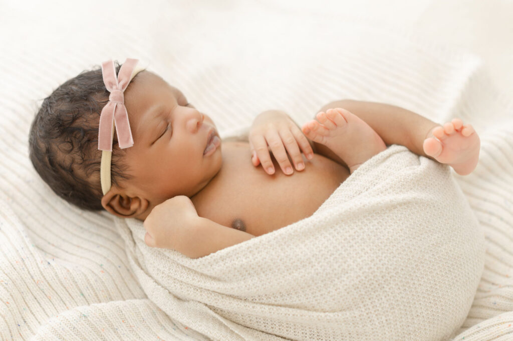 Newborn black baby with light pink bow on her head laying peacefully sleeping on cream blankets and loosely wrapped up in a cream wrap with hands and legs gently out.