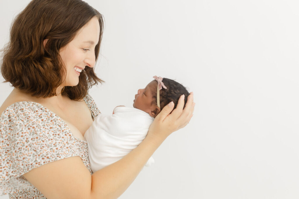 White mom holding black baby. Baby is wrapped in white and sleeping peacefully in moms arms. Mom is smiling very big and looking at baby, admiring her beautiful baby.