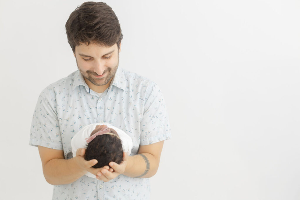 Dad in blue and white floral shirt is holding newborn baby in his arms in front of him and looking down and admiring his new baby.