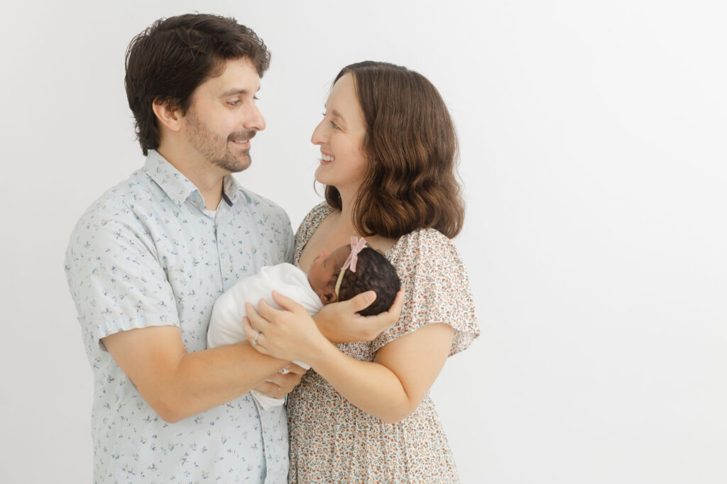 Mom and Dad holding their newborn baby and looking at each other with big smiles on their faces. Studio newborn photography in Portland, Oregon.